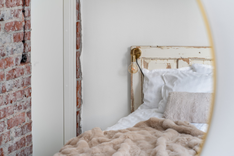 Espejo redondo que refleja una cama blanca junto a una pared de ladrillo visto
