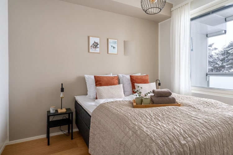 Dormitorio de tonos neutros con cama hecha, cojines naranjas y una gran ventana en esquina