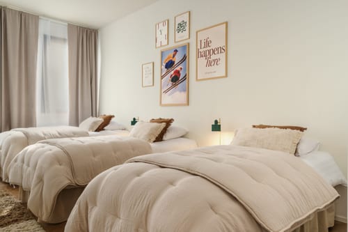 Dormitorio claro con tres camas hechas, edredones beige y láminas enmarcadas en la pared