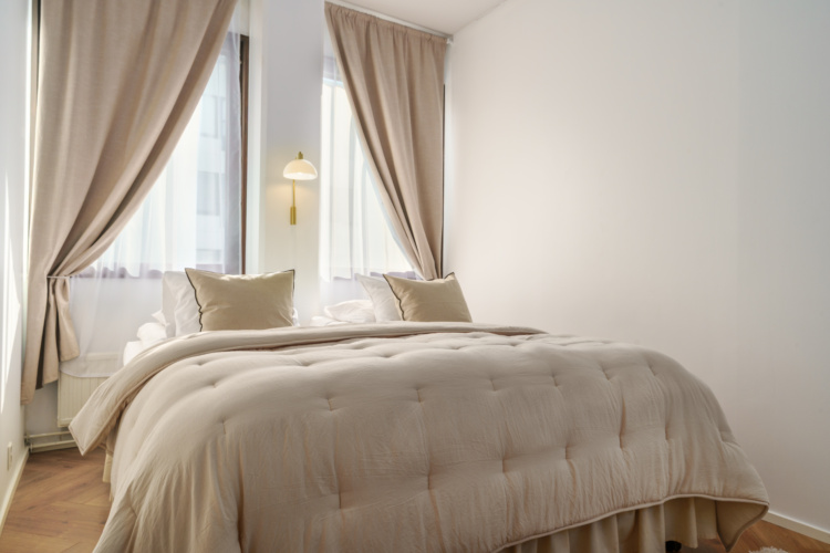 Dormitorio blanco con una cama hecha entre cortinas translúcidas y ventanas altas