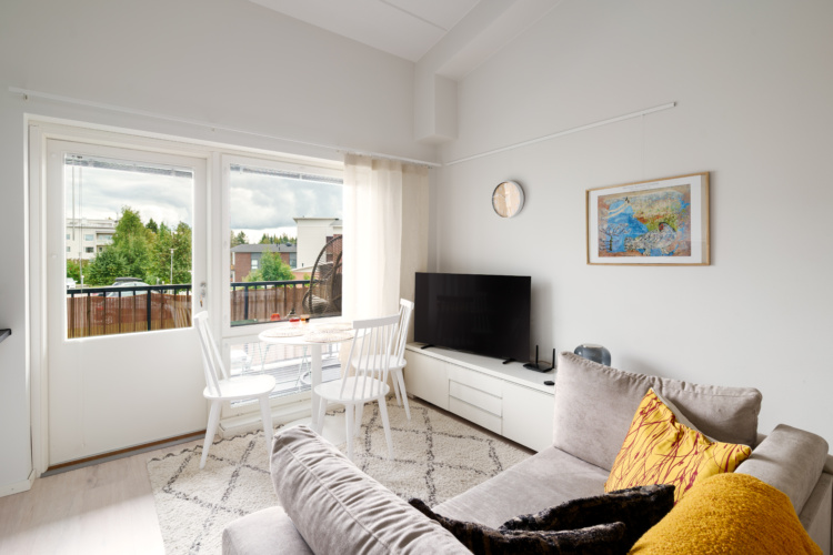 Sala luminosa con sofá gris, juego de comedor blanco, televisor y puertas al balcón