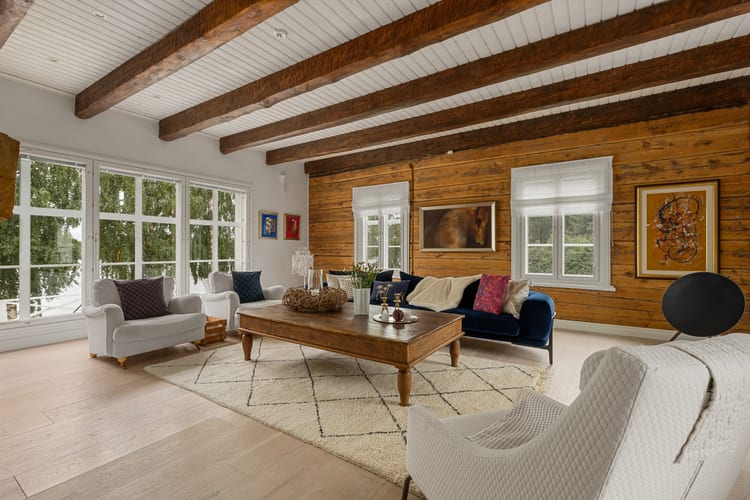 Sala luminosa de una casa de troncos con sofá de terciopelo azul, sillones de cuero y suelo de tablones claros