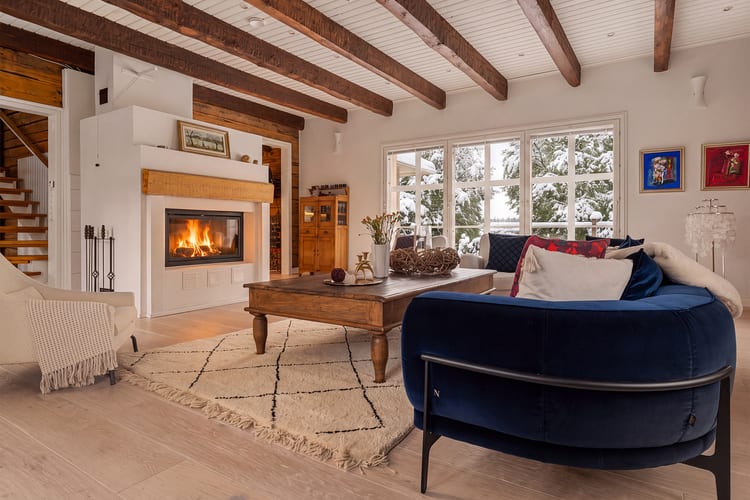 Sala en invierno con fuego encendido en la chimenea, sofá de terciopelo azul y árboles nevados tras las ventanas