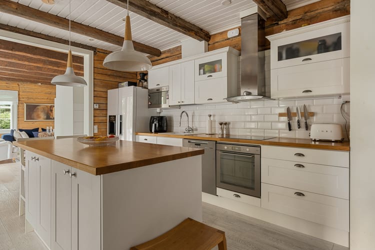 Cocina blanca con isla, lámparas colgantes, taburetes y pared de troncos a la vista