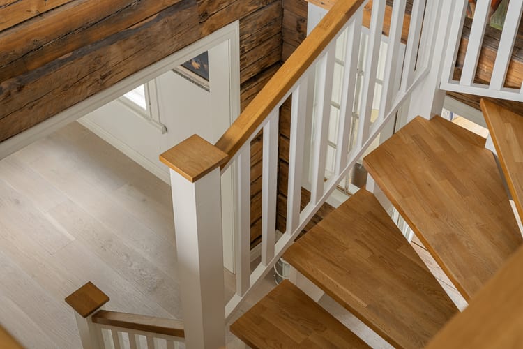 Escalera de roble con barandillas blancas fotografiada desde arriba, con pared de troncos al fondo