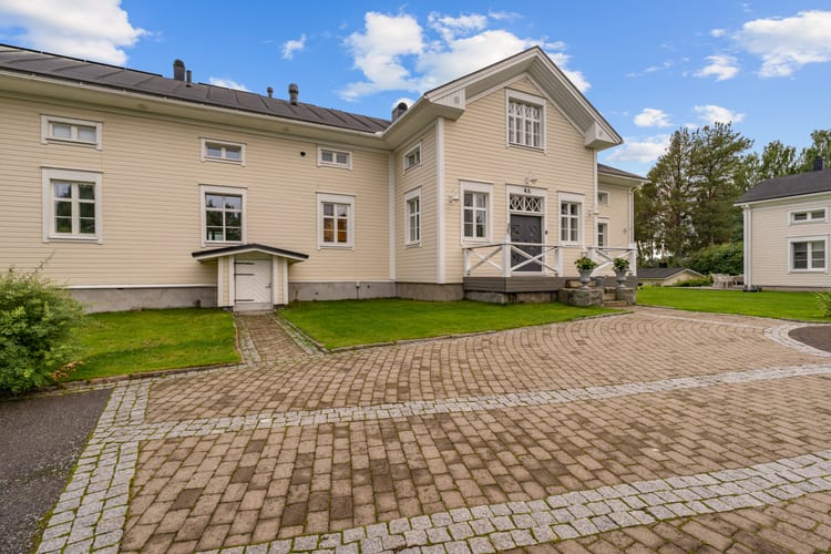 Vaalea kaksikerroksinen päärakennus kesäisenä päivänä, kivetty piha edessä