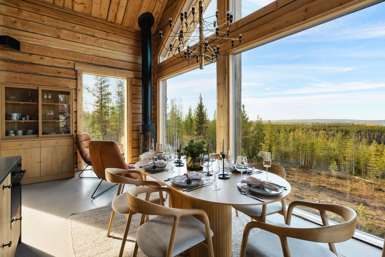 Hirsihuvilan ruokapöytä katettuna panoraamaikkunan edessä, messinkikruunu ja metsämaisema