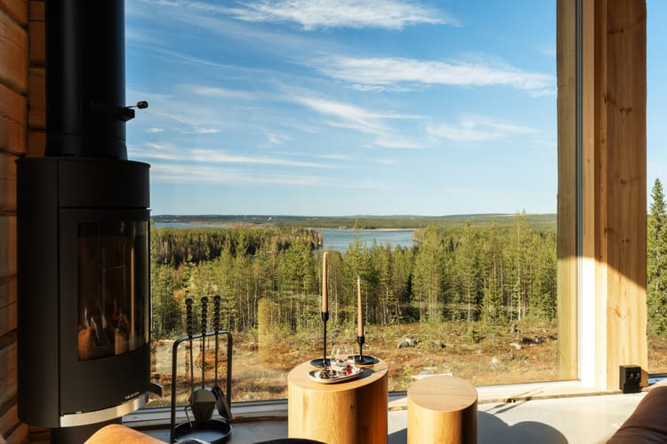 Takassa palaa tuli panoraamaikkunan edessä, kynttilät pöydällä ja järvimaisema ulkona